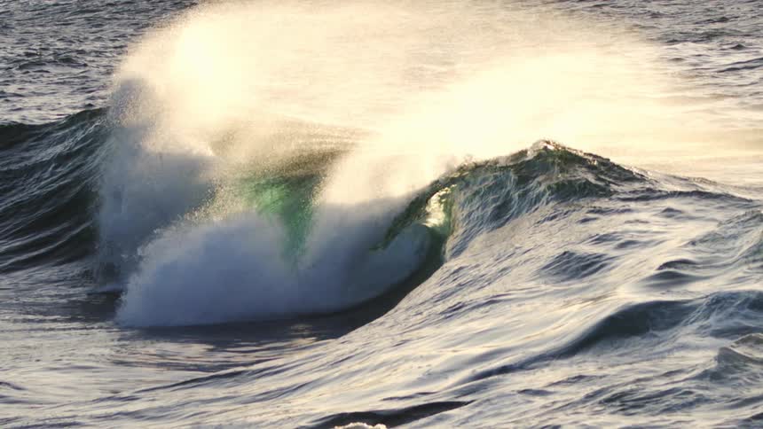 A wave crests and breaks sending up a spray of white foam that catches the sunlight amidst a turbulent sea