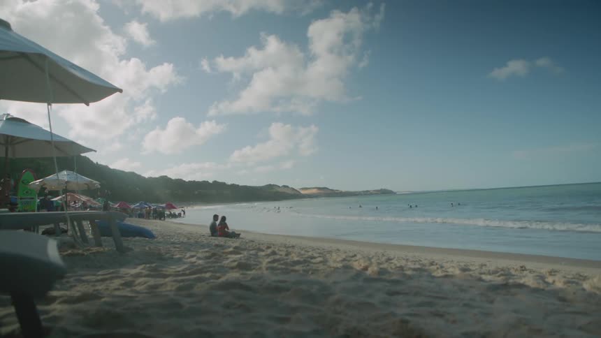 A sunny beach scene captures people relaxing under umbrellas on the sandy shore with the ocean stretching into the distance under a partly cloudy sky