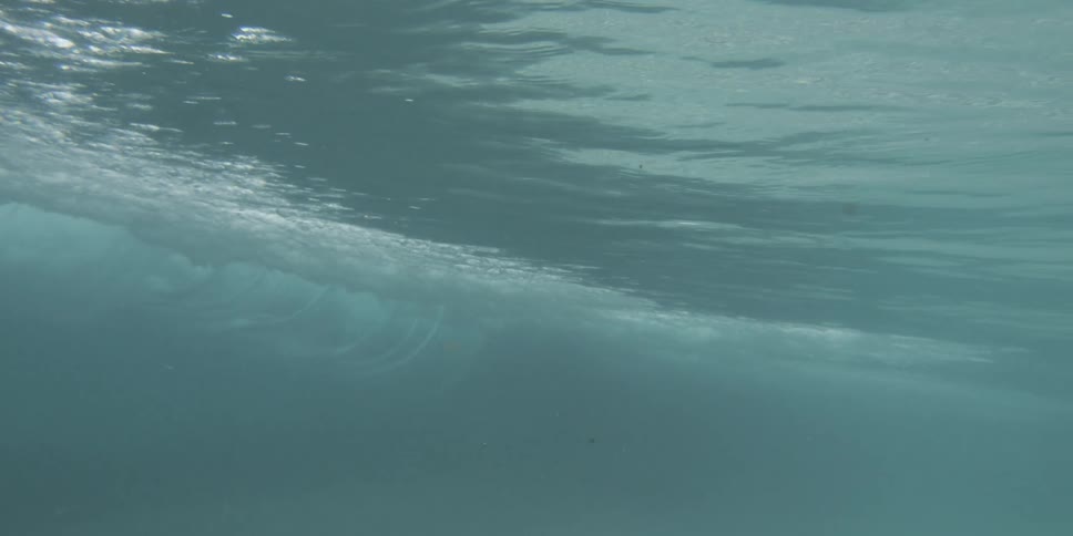 An underwater view of a vast blue ocean shows a wave rippling overhead with bubbles rising in the surface