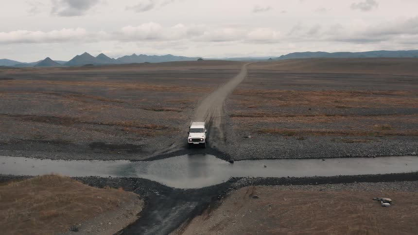A lone white vehicle drives down a narrow road stretching across a vast barren landscape under an overcast sky