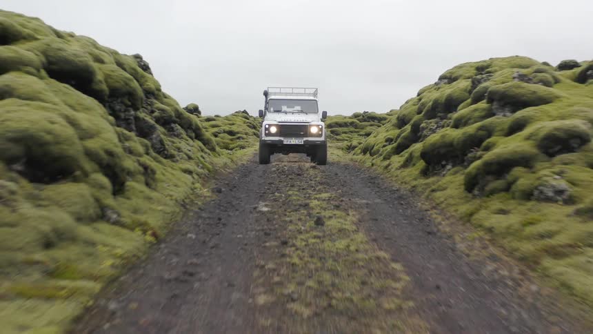 A white land rover defender drives along a dirt track through a mossy rocky landscape under an overcast sky