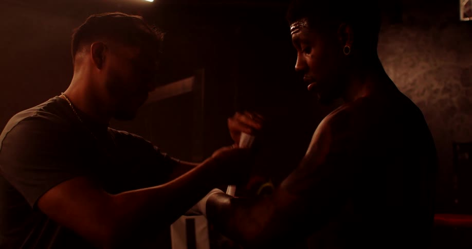 In a dimly lit setting one man carefully wraps another mans hand with white athletic tape preparing for a boxing match