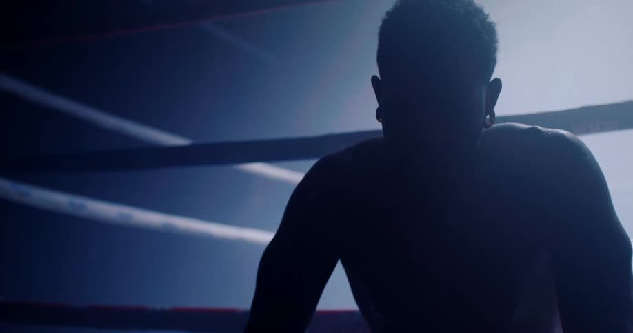 A silhouetted figure of a boxer with an ear gauge sits in a dimly lit boxing ring