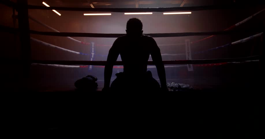 Male boxer silhouetted by flashing lights sits at the edge of the boxing ring and wipes his sweat