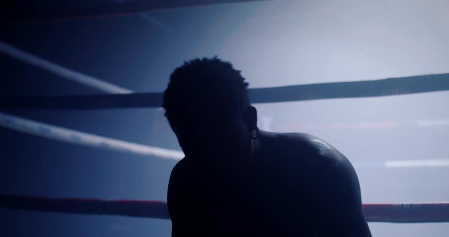 Silhouetted against a bright light a boxer catches his breath and slowly straightens up within the ropes of a boxing ring