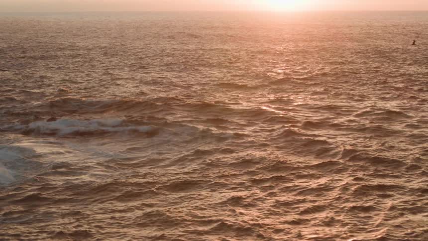 A sunset view captures the oceans surface reflecting the warm light with waves crashing against rocky outcroppings in the foreground