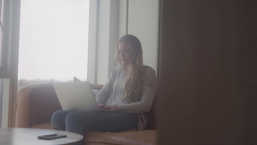 A woman with long blonde hair sits on a couch working on a laptop in a brightly lit room with a partially obscured view of a doorway in the foreground