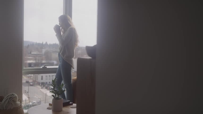A woman with long blonde hair wearing jeans and a light sweater stands by a large window overlooking a cityscape holding a mug to her lips