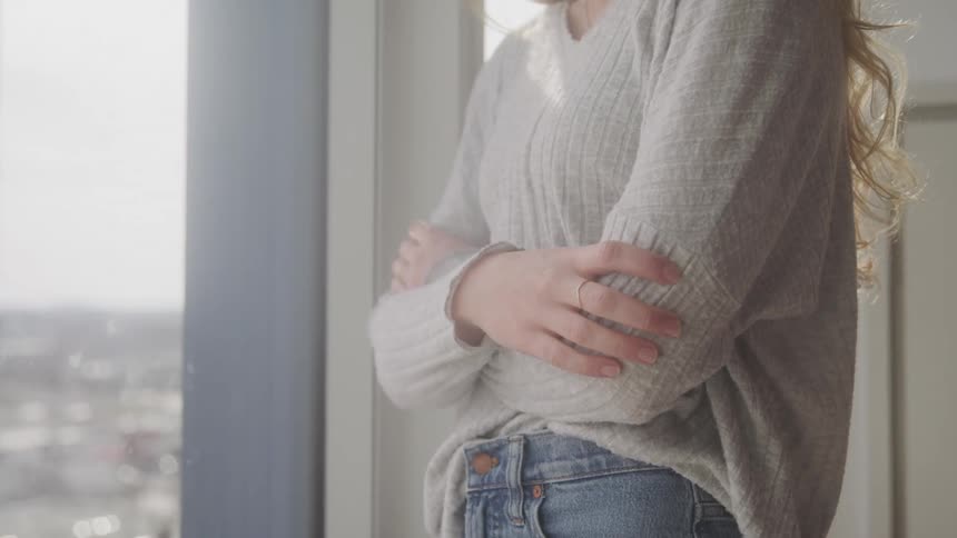 Blonde woman wearing a sweater and jeans stands looking out the window with her arms crossed