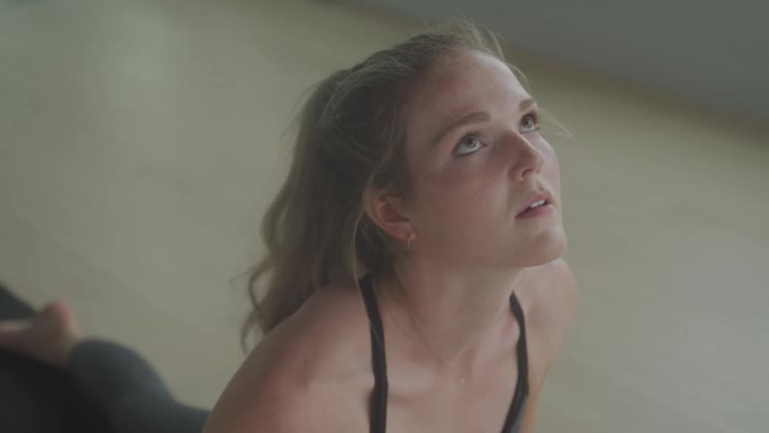 A woman with light hair wearing a black sports bra looking upwards with a contemplative expression as she performs a yoga pose on the floor