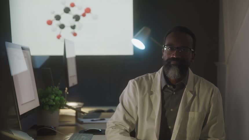A person wearing a lab coat and glasses sits in front of a computer and a large monitor displaying a molecular structure