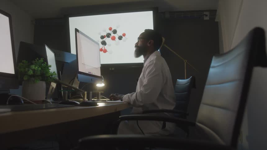 Man wearing a white lab coat works on a computer while the large screen in the background displays the diagram of a molecule