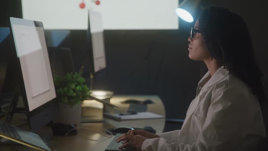 A young woman wearing a white lab coat works at a computer station in a dimly lit laboratory setting
