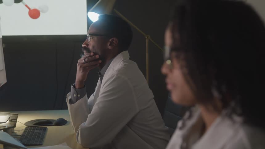 Two individuals in lab coats work at computer stations in a dimly lit room with a molecular structure projected on a screen in the background