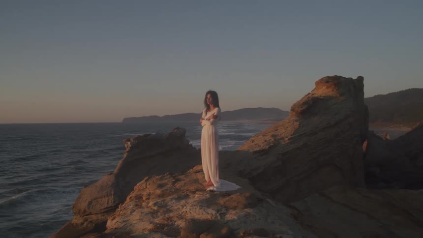 A woman in a white dress stands barefoot on a rocky cliff overlooking the ocean at dusk