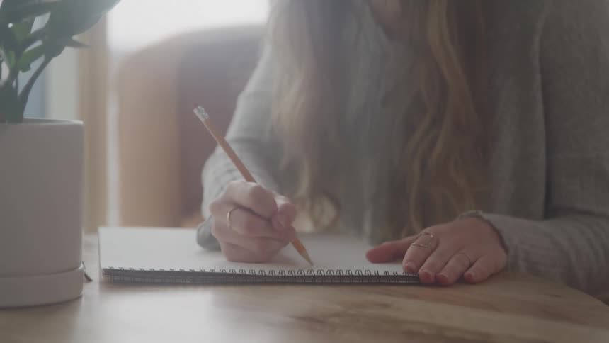 A person writing in a notebook with a pencil beside a potted plant