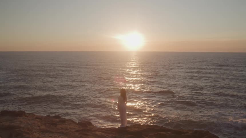 A person stands on a rocky outcrop facing the ocean during a vibrant sunset