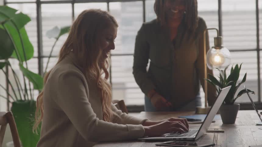 A woman with long blonde hair works on a laptop at a desk while another person with long dark hair stands behind her slightly out of focus in a brightly lit office space with plants and a vintagestyle lamp