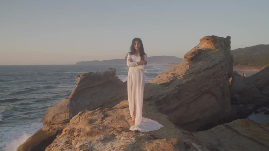 A woman in a long white dress stands on the shore