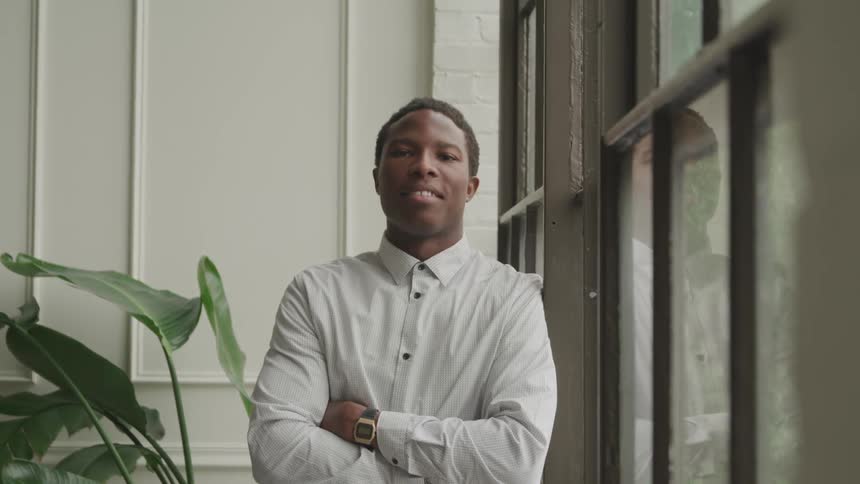 A young man with a warm smile wearing a light gray shirt and a gold watch leans casually against a window frame in a room with white paneled walls and a large potted plant