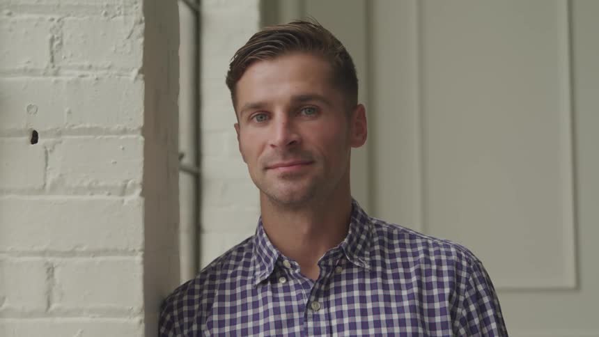 A smiling man in a plaid shirt reclines against a white brick wall looking directly at the camera