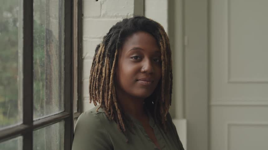 A woman with dreadlocks looking slightly to the side with a gentle smile positioned near a window with a view of greenery