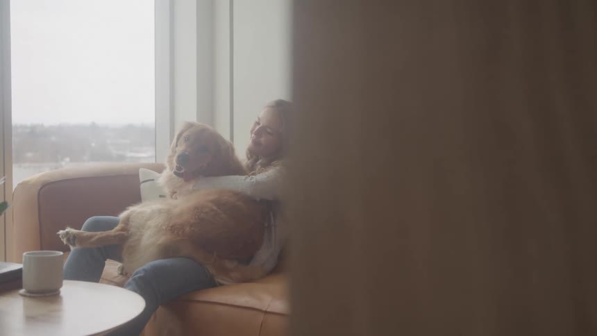 A woman with long blonde hair sits on a couch embracing a golden retriever dog in a bright modern room with a large window overlooking a cityscape