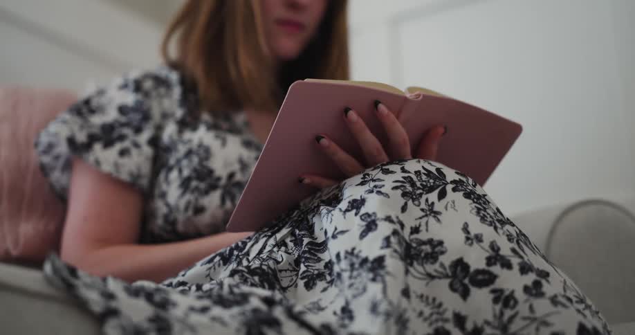 Young woman in a black and white floral dress sitting on couch reading a book