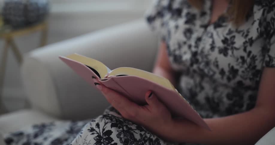A woman in a floral dress sits in a comfortable chair at home reading a pink book near a window in a bright room