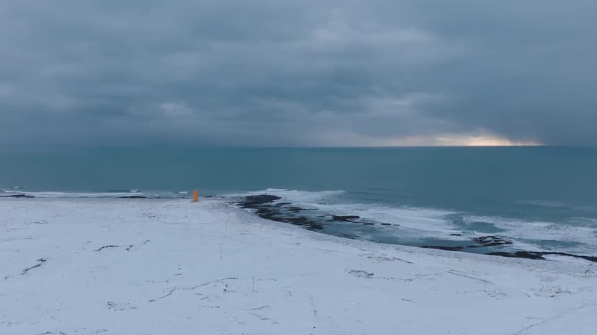 A small orange lighthouse stands on a snowy shoreline with a turquoise sea under a cloudy sky with a sliver of distant sunlight