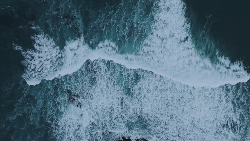 An aerial view of the swirling patterns of frothy waves crashing against a dark rocky shoreline creating a dynamic texture across the oceans surface