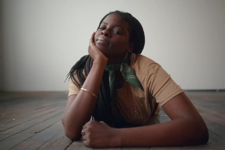 A young woman with dark braids and a tan t shirt poses on a wooden floor leaning on her hand with a thoughtful expression against a plain light colored wall