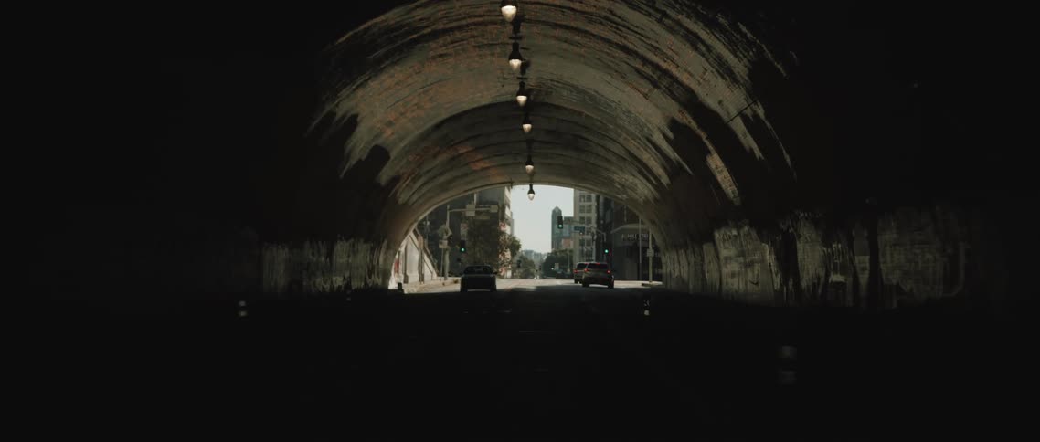 A few cars are driving out of a dimly lit tunnel into the bright daylight of a quiet city street