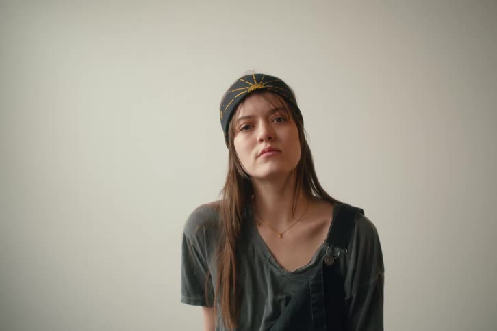 A young woman with long brown hair and a patterned headband looks directly at the camera with a neutral expression against a plain light colored background