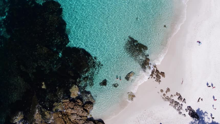 Two people swimming in the crystal clear turquoise water of a shallow cove bordered by a white sandy beach and dark rocky outcrops