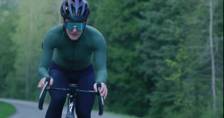 A cyclist wearing a helmet and sunglasses rides along an asphalt path with a blurred forest in the background