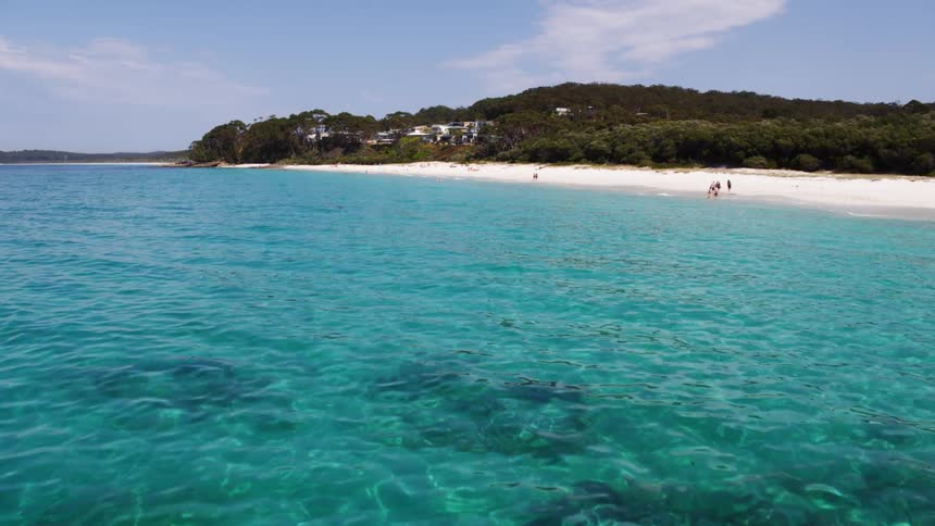 A turquoise ocean leading to a white sandy beach where a few small figures are visible with a forested hillside in the background under a partly cloudy sky
