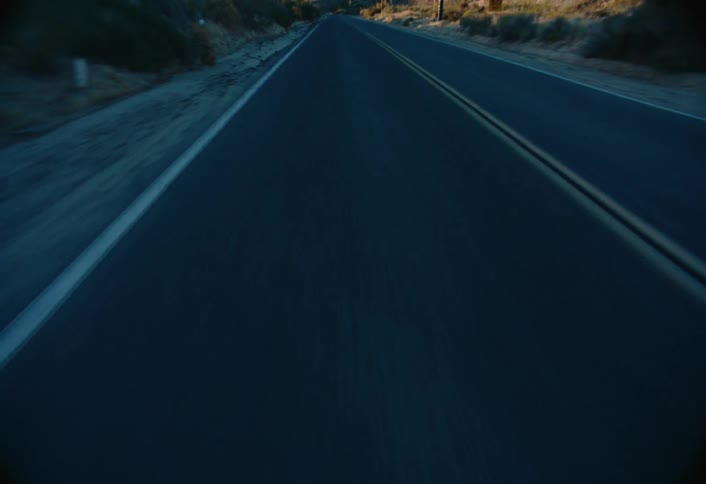 A paved road with double yellow lines stretches through a desert landscape with mountains under a clear sky