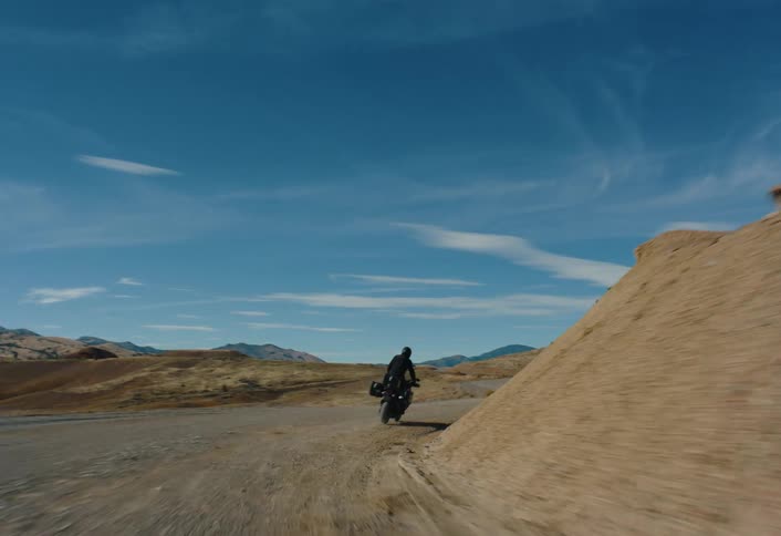 A person rides a motorcycle loaded with gear along a winding dirt road through a barren hilly landscape under a bright sky