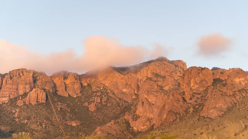A rugged mountain range bathed in the warm light of sunset with the rocky peaks and cliffs illuminated against a soft pastel colored sky
