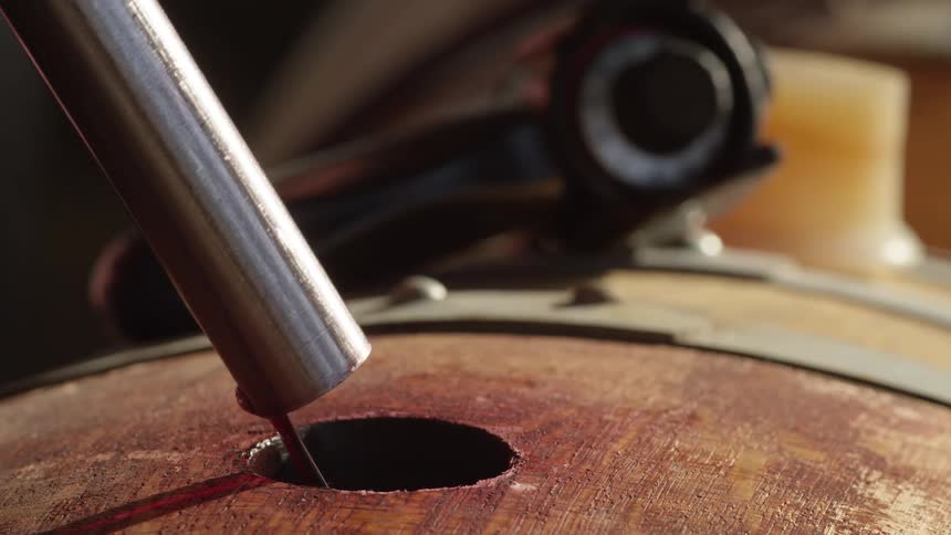 A closeup shows a metal pipe pouring foaming red liquid into a wooden barrel through a hole until it fills completely