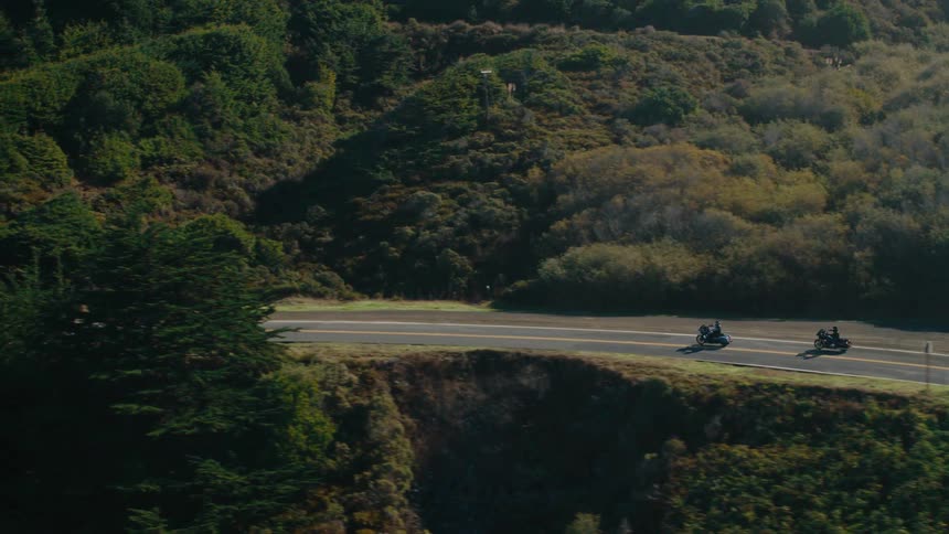 Two motorcycles travel along a winding coastal highway carved into a steep vegetationcovered hillside