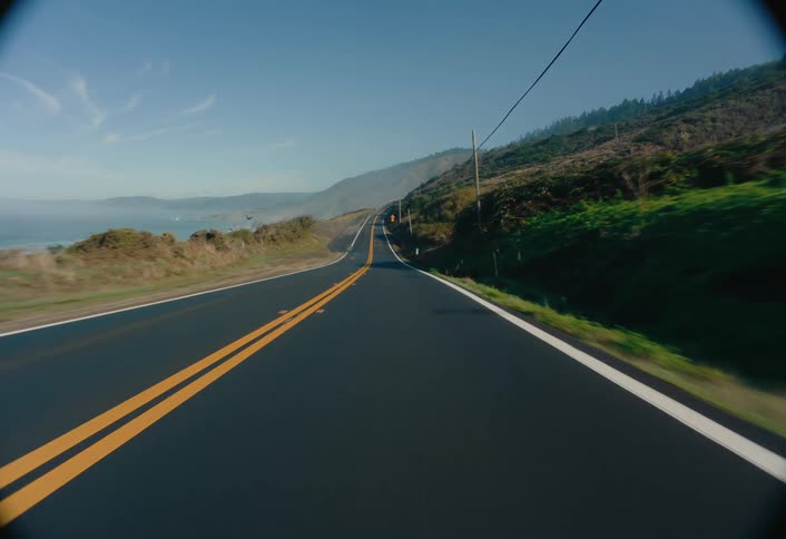 A moving vehicle captures an asphalt road stretching into the distance under a clear blue sky with a hillside on one side and a glimpse of the ocean on the other