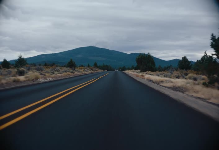 A moving vehicle captures an asphalt road stretching into the distance under a cloudy sky with dry shrubs lining the sides and a mountain visible in the background
