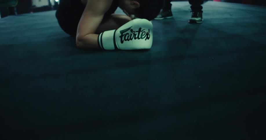 A boxer wearing gloves lies on the floor inside a boxing ring slamming her glove against the mat in frustration while another person stands close by