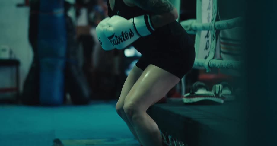 A female boxer wearing white gloves sits on the side of a ring preparing for practice then stands up and walks away