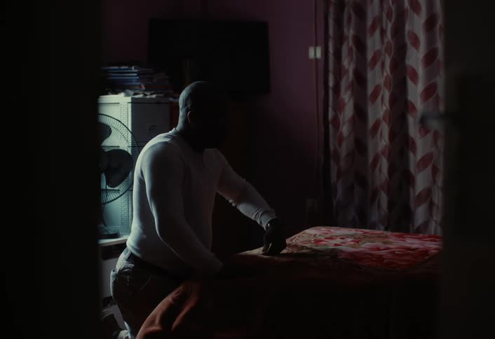 A person is kneeling beside a bed in a dimly lit room with a fan stacks of books and a television in the background