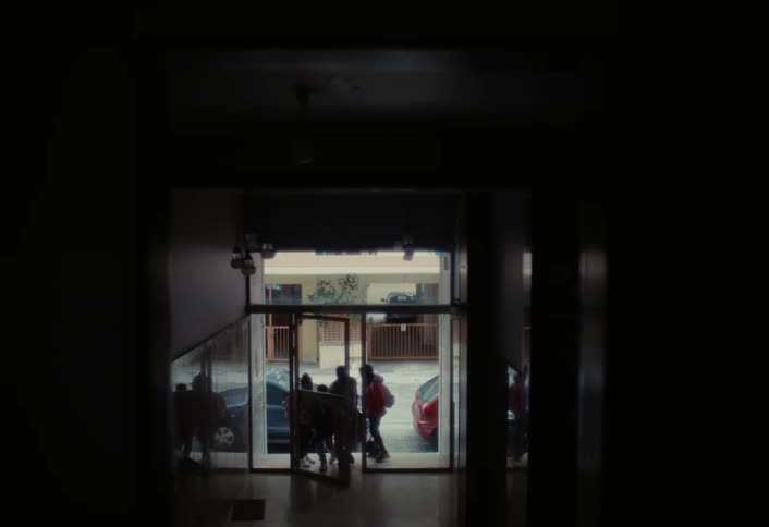 A family steps through a glass door from the brightly lit street into the shadowy hallway of a residential building