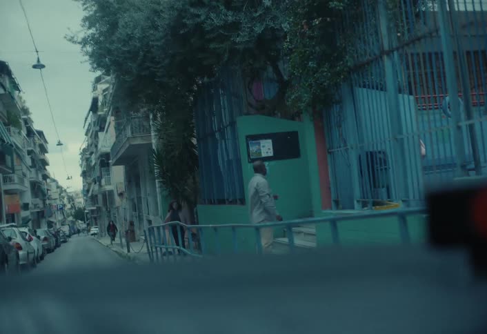From a vehicle a father is seen picking up his daughter at school with a cityscape and an overcast sky in the background