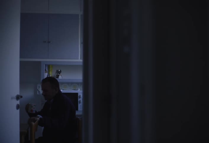 A dimly lit interior shot shows a person sitting at a table in the kitchen drinking from a mug